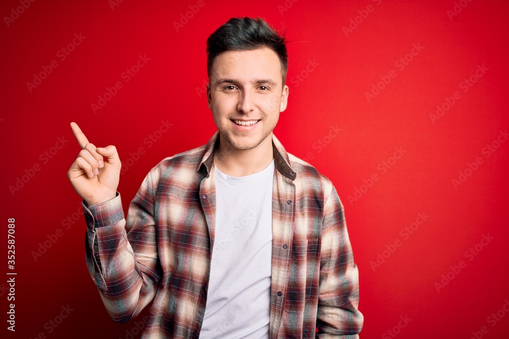 Young handsome caucasian man wearing casual modern shirt over red isolated background with a big smile on face, pointing with hand finger to the side looking at the camera. Young handsome caucasian man wearing casual modern shirt over red isolated background with a big smile on face, pointing with hand finger to the side looking at the camera.