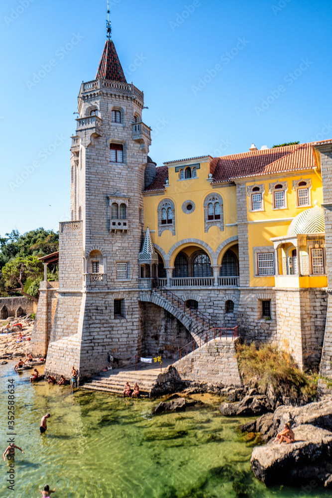 The Palácio dos Condes de Castro Guimarães illuminated by the sun with tourists bathing in the water at the bottom of the steps.