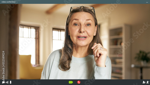 Portrait of casually dressed energetic elderly woman with loose gray hair posing in stylish apartment interior looking at camera with mouth opened, speaking via virtual chat, making webcam video call