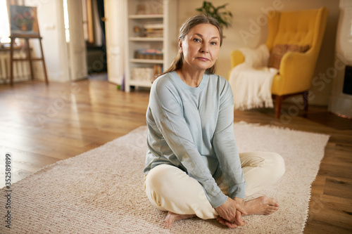 Billede på lærred Attractive gray haired retired woman sitting barefooted on carpet at home, keeping legs crossed, stretching thighs, doing butterfly yoga exercise