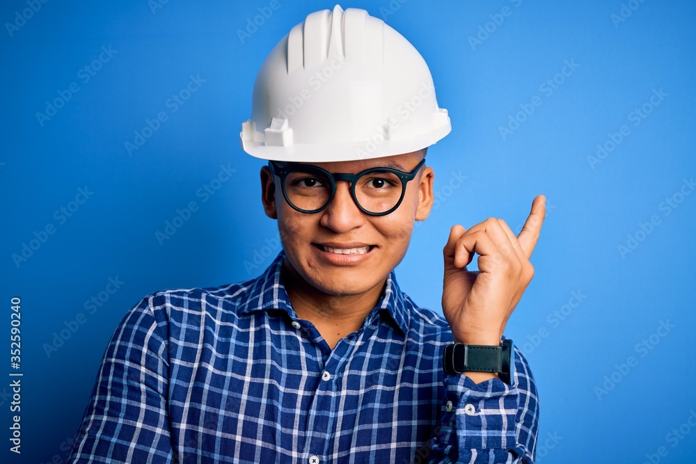 Young handsome engineer latin man wearing safety helmet over isolated blue background with a big smile on face, pointing with hand and finger to the side looking at the camera. Young handsome engineer latin man wearing safety helmet over isolated blue background with a big smile on face, pointing with hand and finger to the side looking at the camera.