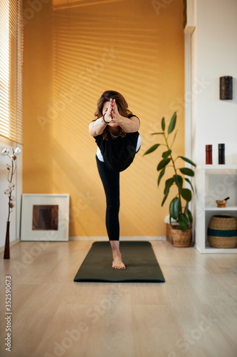 Attractive fit smiling middle aged yogi woman standing on mat in Lord of the Dance yoga pose at home.