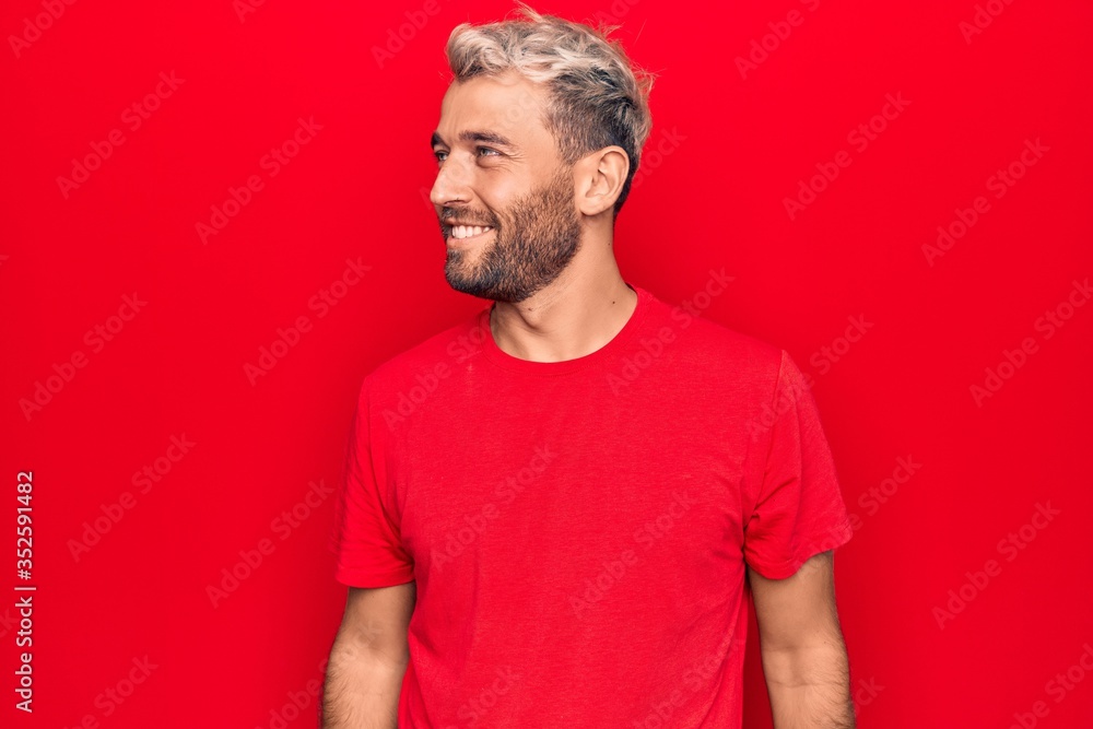 Young handsome blond man wearing casual red t-shirt standing over isolated red background looking to side, relax profile pose with natural face and confident smile. Young handsome blond man wearing casual red t-shirt standing over isolated red background looking to side, relax profile pose with natural face and confident smile.