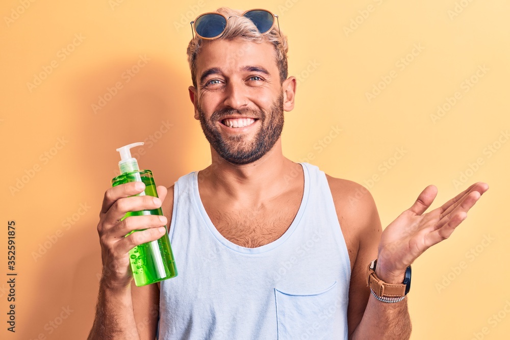 Handsome blond man on vacation holding bottle of aloe vera cream over yellow background celebrating achievement with happy smile and winner expression with raised hand Handsome blond man on vacation holding bottle of aloe vera cream over yellow background celebrating achievement with happy smile and winner expression with raised hand