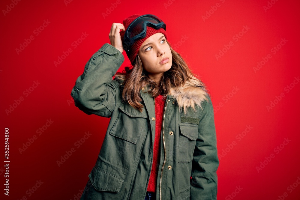 Young blonde girl wearing ski glasses and winter coat for ski weather over red background confuse and wondering about question. Uncertain with doubt, thinking with hand on head. Pensive concept. Young blonde girl wearing ski glasses and winter coat for ski weather over red background confuse and wondering about question. Uncertain with doubt, thinking with hand on head. Pensive concept.