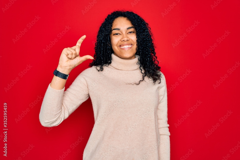 Young african american curly woman wearing casual turtleneck sweater over red background smiling and confident gesturing with hand doing small size sign with fingers looking and the camera. Measure Young african american curly woman wearing casual turtleneck sweater over red background smiling and confident gesturing with hand doing small size sign with fingers looking and the camera. Measure