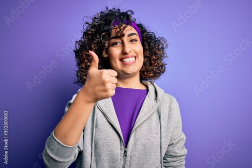Beautiful curly arab sportswoman doing sport wearing sportswear over purple background doing happy thumbs up gesture with hand. Approving expression looking at the camera showing success.