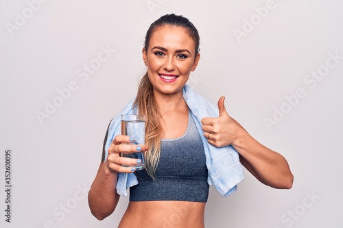 Beautiful blonde sporty woman using towel drinking bottle of water over white background smiling happy and positive, thumb up doing excellent and approval sign