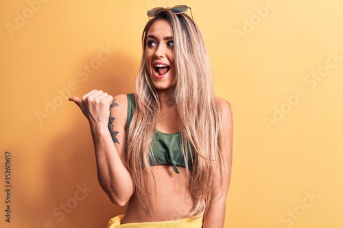 Young beautiful blonde woman wearing bikini and sunglasses standing over yellow background pointing thumb up to the side smiling happy with open mouth