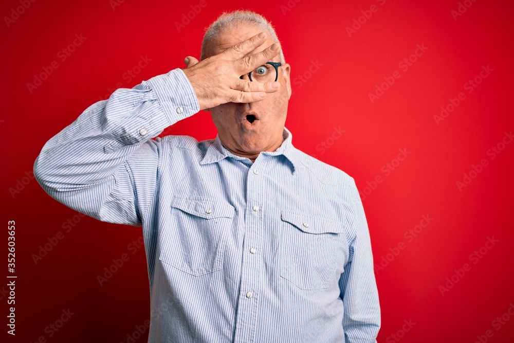 Middle age handsome hoary man wearing casual striped shirt and glasses over red background peeking in shock covering face and eyes with hand, looking through fingers with embarrassed expression. Middle age handsome hoary man wearing casual striped shirt and glasses over red background peeking in shock covering face and eyes with hand, looking through fingers with embarrassed expression.