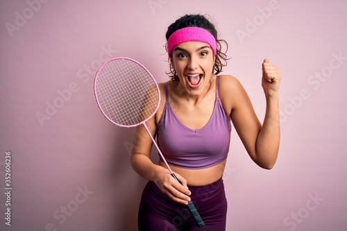 Beautiful sportswoman with curly hair wearing sportswear playing badminton uning racket screaming proud and celebrating victory and success very excited, cheering emotion photo