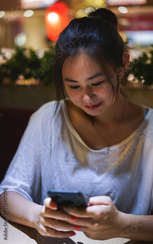 Beautiful cute happy smiling Asian Chinese girl using phone at the restaurant, ordering food online, gadget addiction, no talking with friends, life online concept