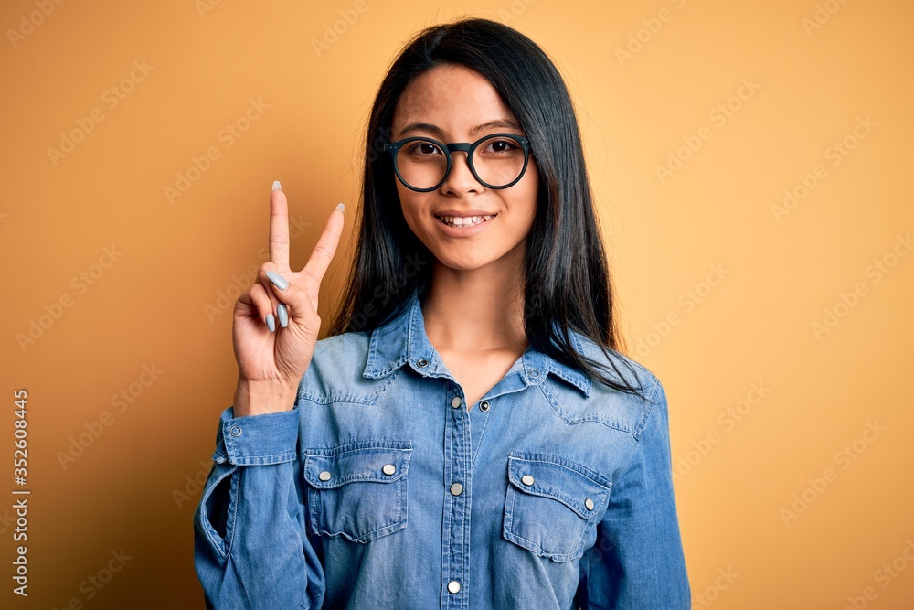 Young beautiful chinese woman wearing casual denim shirt over isolated yellow background smiling looking to the camera showing fingers doing victory sign. Number two. Young beautiful chinese woman wearing casual denim shirt over isolated yellow background smiling looking to the camera showing fingers doing victory sign. Number two.