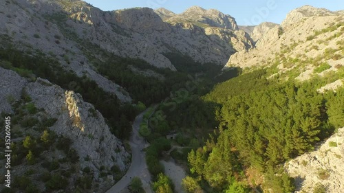 Aerial shot of road by trees in valley with rocky mountains against sky, drone flying forward over green forest on sunny day - Pag, Croatia photo