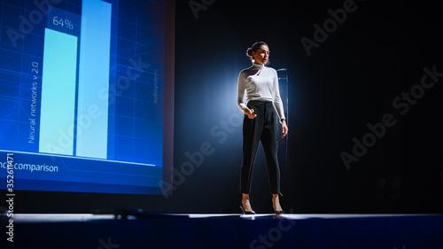 On-Stage Successful Female Speaker Presents Technological Product, Uses Remote Control for Presentation, Showing Infographics, Statistics Animation on Screen. Live Event / Device Release. © Gorodenkoff