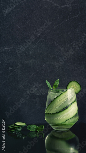 View of drinking glass with cucumber tonic gin cocktail with fresh mint on glossy black table and background refreshing long drink summer recipe