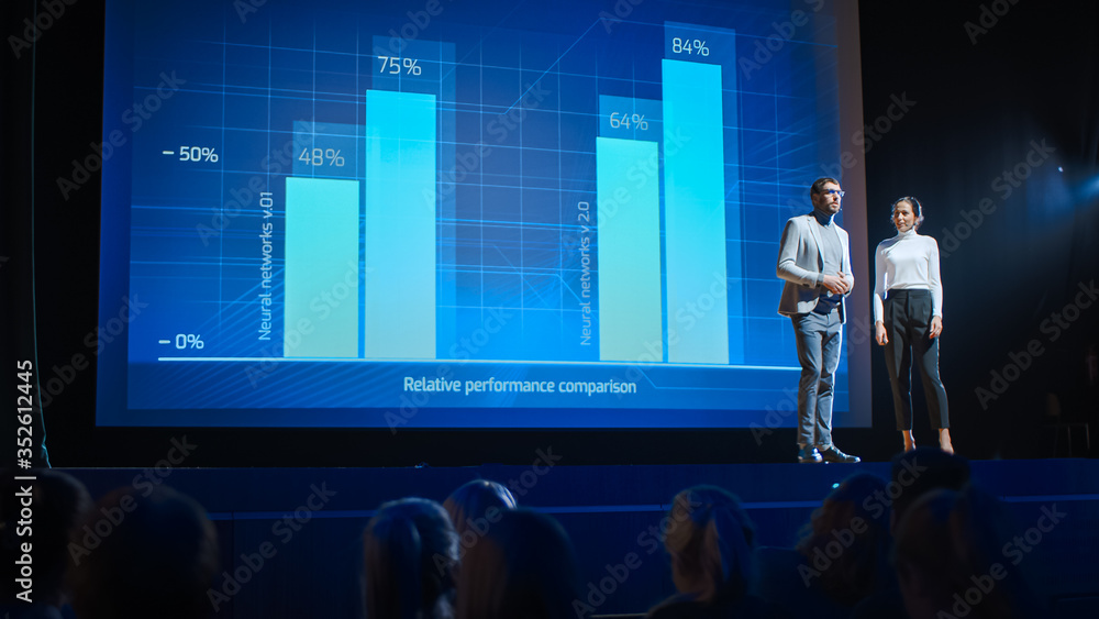 © Gorodenkoff - On Stage, Successful Female Executive and Male Manager Present New Product, Show Infographics, Statistics on Big Screen, Talk About Growth. Live Event, Tech Startup, Business Conference