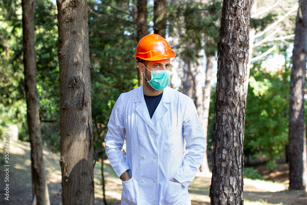 Ricercatore con caschetto arancione , camice bianco e mascherina protettiva immerso nella natura di un bosco Ricercatore con caschetto arancione , camice bianco e mascherina protettiva immerso nella natura di un bosco
