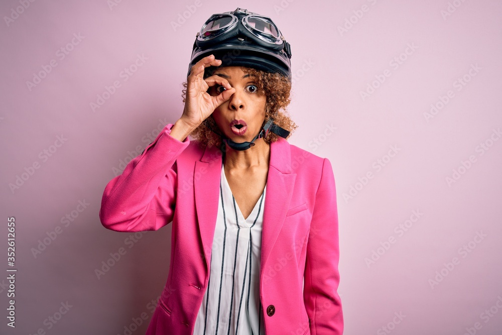 African american motorcyclist woman with curly hair wearing moto helmet over pink background doing ok gesture shocked with surprised face, eye looking through fingers. Unbelieving expression. African american motorcyclist woman with curly hair wearing moto helmet over pink background doing ok gesture shocked with surprised face, eye looking through fingers. Unbelieving expression.