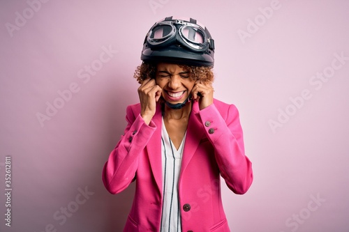 African american motorcyclist woman with curly hair wearing moto helmet over pink background covering ears with fingers with annoyed expression for the noise of loud music. Deaf concept.