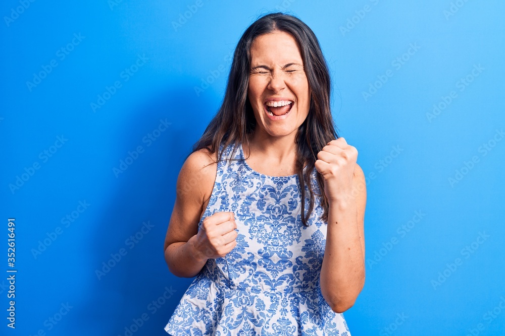 Young beautiful brunette woman wearing casual sleeveless t-shirt over blue background celebrating surprised and amazed for success with arms raised and eyes closed Young beautiful brunette woman wearing casual sleeveless t-shirt over blue background celebrating surprised and amazed for success with arms raised and eyes closed