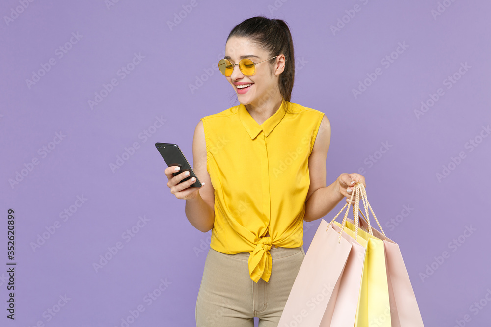 Smiling young brunette woman girl in yellow shirt posing isolated on violet background. People lifestyle concept. Mock up copy space. Hold package bag with purchases after shopping using mobile phone. Smiling young brunette woman girl in yellow shirt posing isolated on violet background. People lifestyle concept. Mock up copy space. Hold package bag with purchases after shopping using mobile phone.