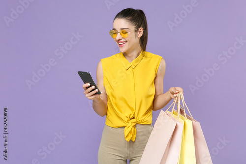Smiling young brunette woman girl in yellow shirt posing isolated on violet background. People lifestyle concept. Mock up copy space. Hold package bag with purchases after shopping using mobile phone.