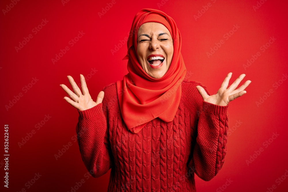 Middle age woman wearing traditional muslim hijab standing over isolated red background celebrating mad and crazy for success with arms raised and closed eyes screaming excited. Winner concept Middle age woman wearing traditional muslim hijab standing over isolated red background celebrating mad and crazy for success with arms raised and closed eyes screaming excited. Winner concept