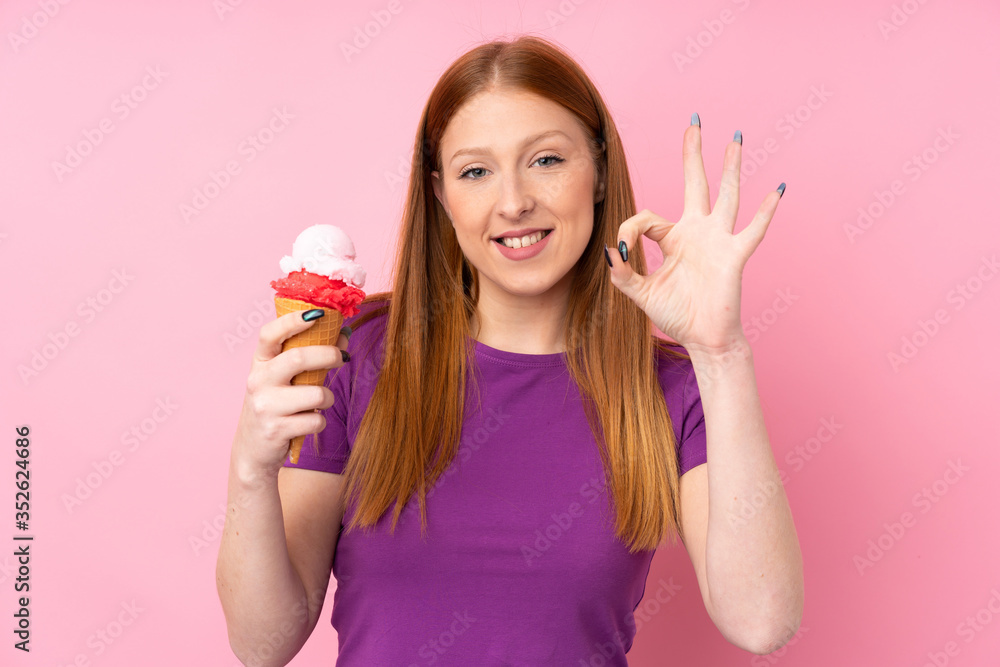 Young redhead woman with a cornet ice cream over isolated pink background showing ok sign with fingers Young redhead woman with a cornet ice cream over isolated pink background showing ok sign with fingers