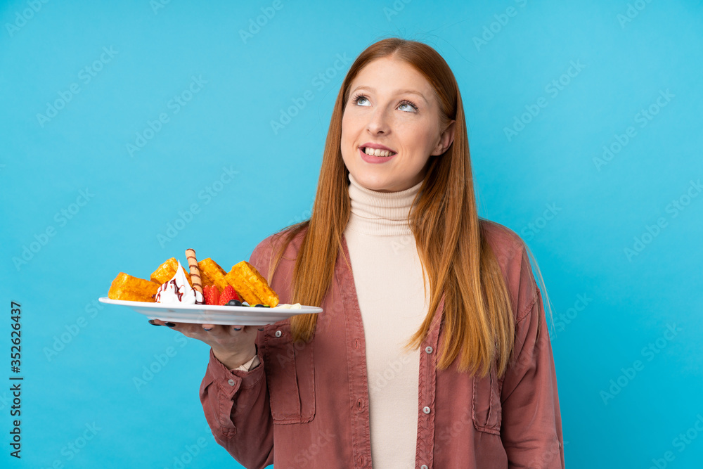 Young redhead woman holding waffles over isolated background looking up while smiling Young redhead woman holding waffles over isolated background looking up while smiling