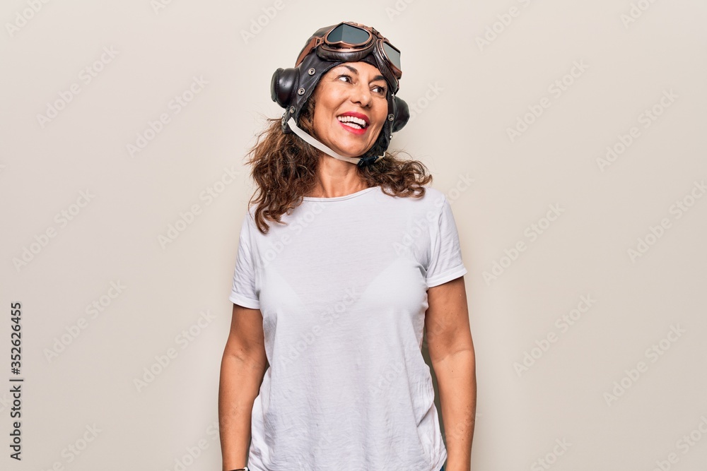 Middle age brunette woman wearing goggles and retro aviator leather hat over white background looking to side, relax profile pose with natural face and confident smile. Middle age brunette woman wearing goggles and retro aviator leather hat over white background looking to side, relax profile pose with natural face and confident smile.