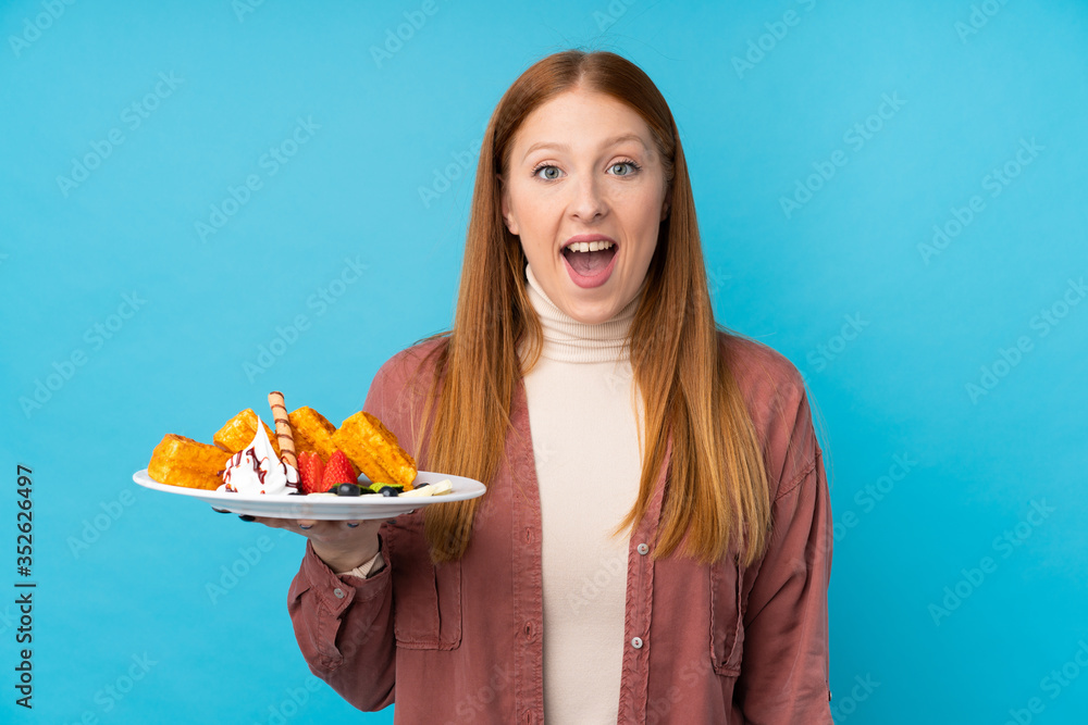 Young redhead woman holding waffles over isolated background with surprise and shocked facial expression Young redhead woman holding waffles over isolated background with surprise and shocked facial expression