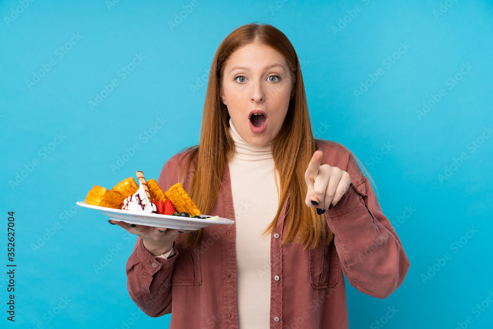 Young redhead woman holding waffles over isolated background surprised and pointing front Young redhead woman holding waffles over isolated background surprised and pointing front