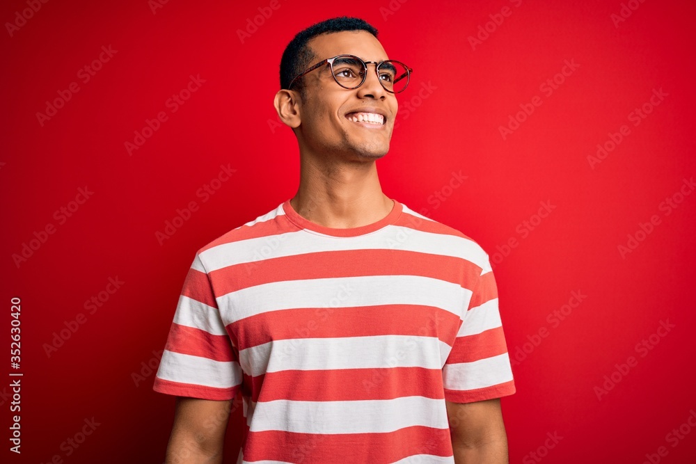 Young handsome african american man wearing casual striped t-shirt and glasses looking away to side with smile on face, natural expression. Laughing confident. Young handsome african american man wearing casual striped t-shirt and glasses looking away to side with smile on face, natural expression. Laughing confident.