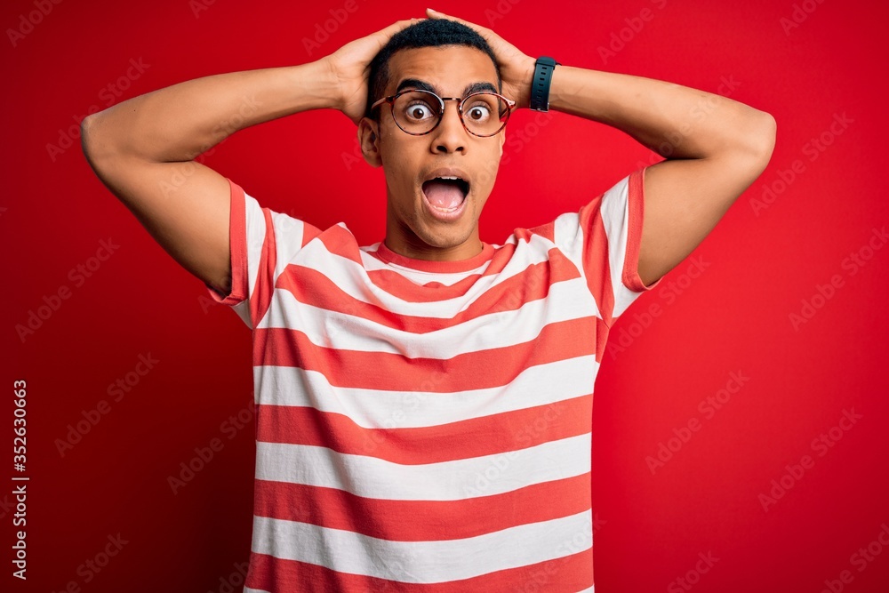 Young handsome african american man wearing casual striped t-shirt and glasses Crazy and scared with hands on head, afraid and surprised of shock with open mouth Young handsome african american man wearing casual striped t-shirt and glasses Crazy and scared with hands on head, afraid and surprised of shock with open mouth