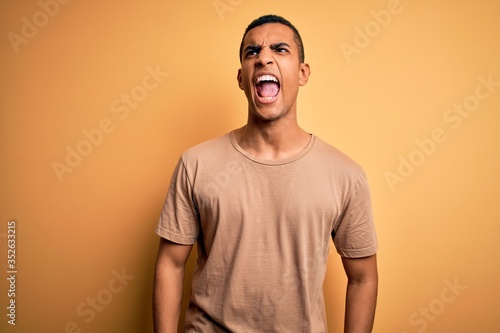 Young handsome african american man wearing casual t-shirt standing over yellow background angry and mad screaming frustrated and furious, shouting with anger. Rage and aggressive concept.