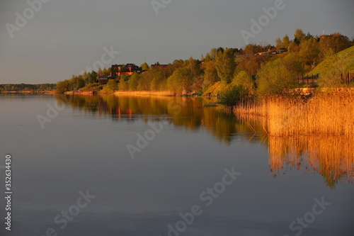 High Bank of the river with houses on top illuminated by a bright orange light at dawn.The mirror image of trees and tall grass on the smooth surface of the water.Panorama of the sleeping coast.Russia