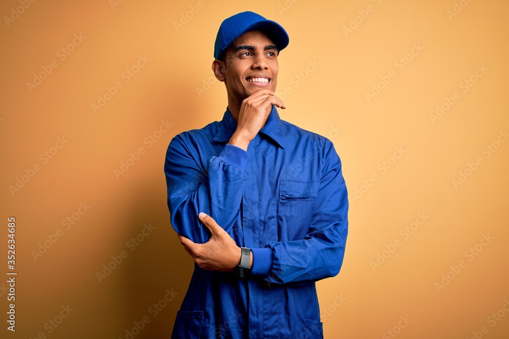 Young african american mechanic man wearing blue uniform and cap over yellow background with hand on chin thinking about question, pensive expression. Smiling with thoughtful face. Doubt concept. Young african american mechanic man wearing blue uniform and cap over yellow background with hand on chin thinking about question, pensive expression. Smiling with thoughtful face. Doubt concept.