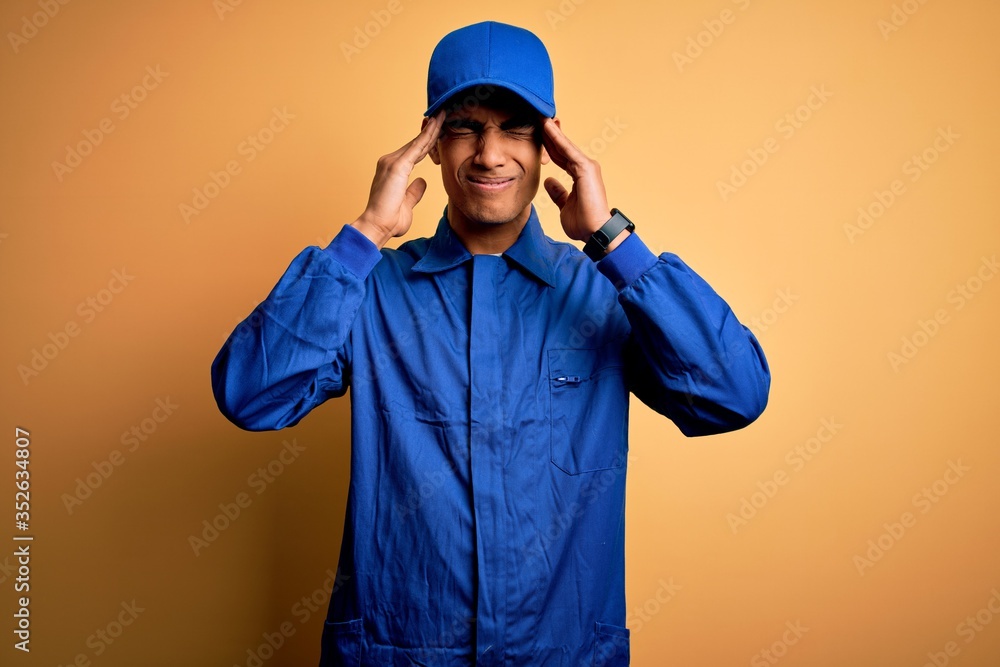 Young african american mechanic man wearing blue uniform and cap over yellow background with hand on head for pain in head because stress. Suffering migraine.