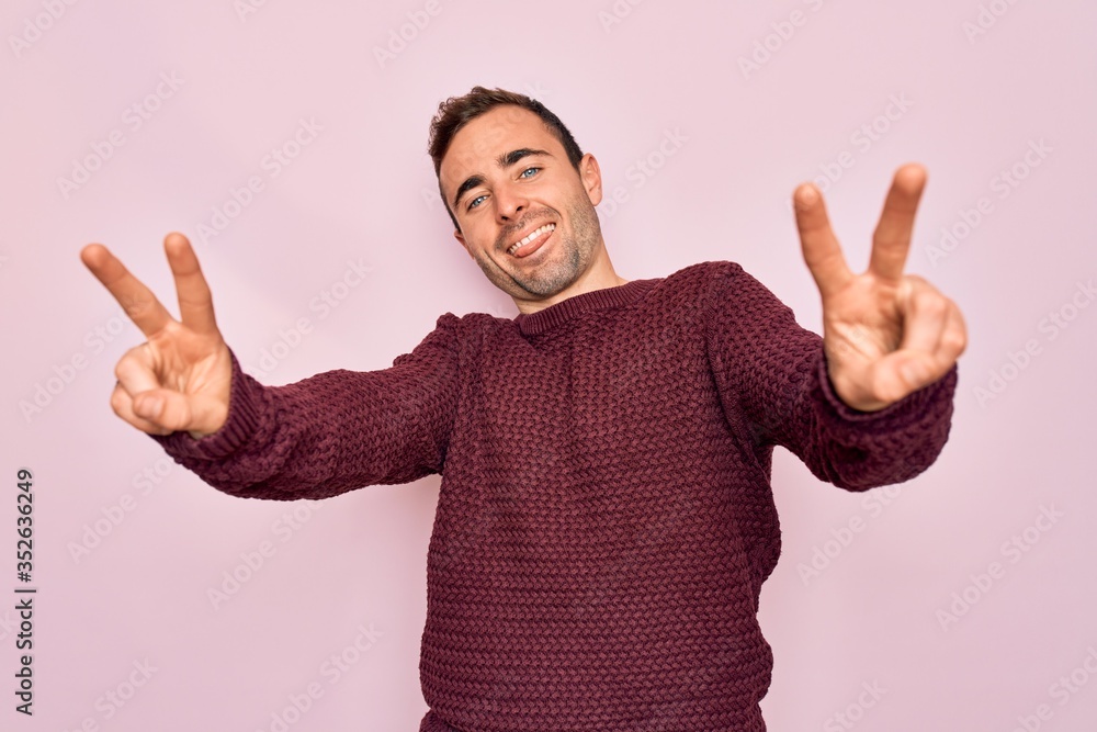 Young handsome man with blue eyes wearing casual sweater standing over pink background smiling with tongue out showing fingers of both hands doing victory sign. Number two. Young handsome man with blue eyes wearing casual sweater standing over pink background smiling with tongue out showing fingers of both hands doing victory sign. Number two.