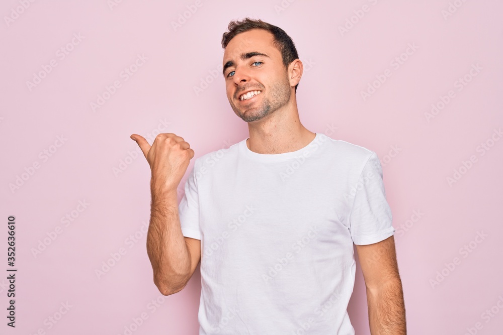 Handsome man with blue eyes wearing casual white t-shirt standing over pink background smiling with happy face looking and pointing to the side with thumb up. Handsome man with blue eyes wearing casual white t-shirt standing over pink background smiling with happy face looking and pointing to the side with thumb up.