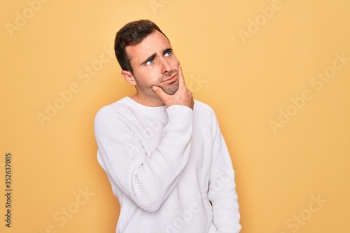 Young handsome man with blue eyes wearing casual sweater standing over yellow background Thinking worried about a question, concerned and nervous with hand on chin