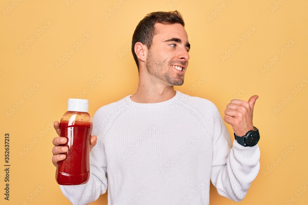 Young handsome man holding bottle of ketchup sauce condiment over yellow background pointing and showing with thumb up to the side with happy face smiling Young handsome man holding bottle of ketchup sauce condiment over yellow background pointing and showing with thumb up to the side with happy face smiling