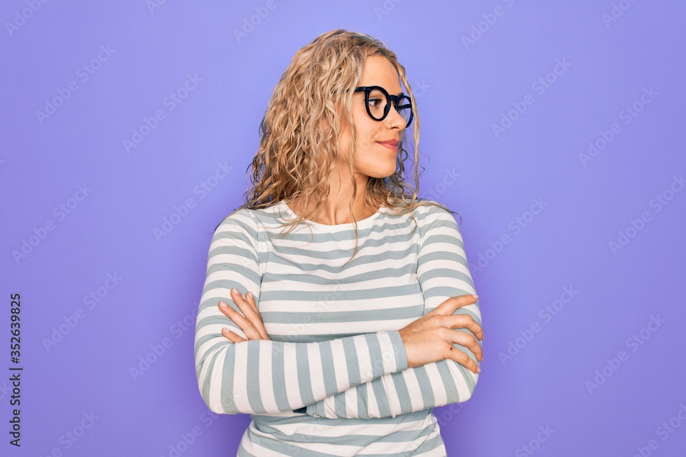 Fototapeta premium Beautiful blonde woman wearing casual striped t-shirt and glasses over purple background looking to the side with arms crossed convinced and confident