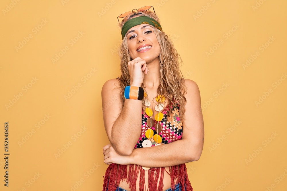 Beautiful blonde hippie woman wearing sunglasses and accessories over yellow background looking confident at the camera with smile with crossed arms and hand raised on chin. Thinking positive. Beautiful blonde hippie woman wearing sunglasses and accessories over yellow background looking confident at the camera with smile with crossed arms and hand raised on chin. Thinking positive.