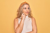 Beautiful blonde woman wearing casual t-shirt and diadem over isolated yellow background with hand on chin thinking about question, pensive expression. Smiling with thoughtful face. Doubt concept.