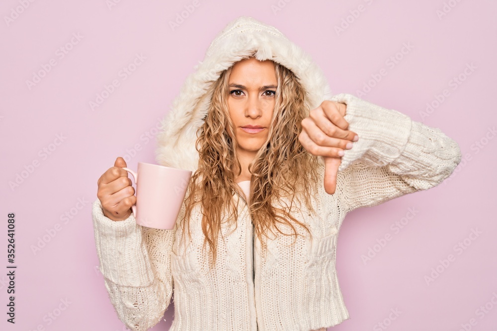 Young beautiful blonde woman wearing casual sweater with hood drinking cup of coffee with angry face, negative sign showing dislike with thumbs down, rejection concept Young beautiful blonde woman wearing casual sweater with hood drinking cup of coffee with angry face, negative sign showing dislike with thumbs down, rejection concept