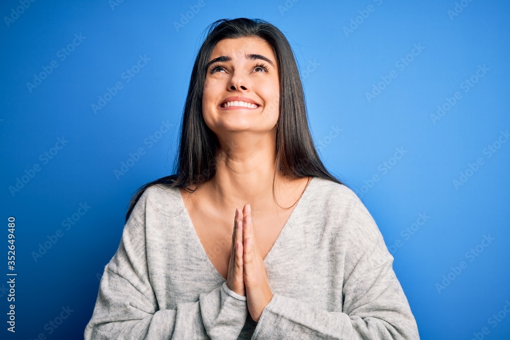Obraz premium Young beautiful brunette woman wearing casual sweater standing over blue background begging and praying with hands together with hope expression on face very emotional and worried. Begging.