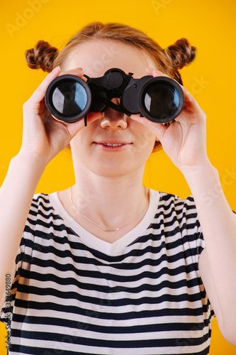 Cute ginger woman in a striped dress looking in binoculars. Close up. Her hair in two small knot buns. Over orange background.