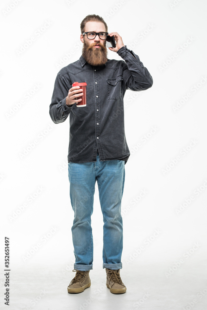 Handsome bearded man with stylish hair beard and mustache talking on mobile phone holding cup or mug drinking tea or coffee on white background Handsome bearded man with stylish hair beard and mustache talking on mobile phone holding cup or mug drinking tea or coffee on white background